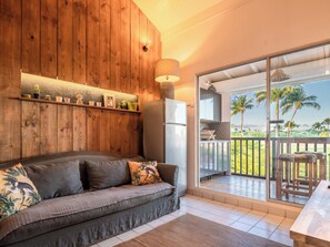 Una habitación acogedora con pared de madera, un sofá con cojines, un refrigerador y un amplio ventanal corredera que da a un balcón con vistas a palmeras.