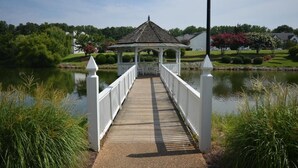 A wooden bridge leading to a gazebo over a calm lake, surrounded by greenery and houses.