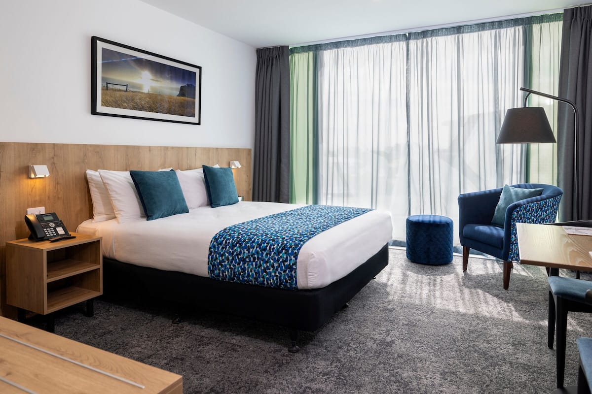 A hotel room with a bed, bedside table, lamp, chair, and a view of the outdoors through large windows.