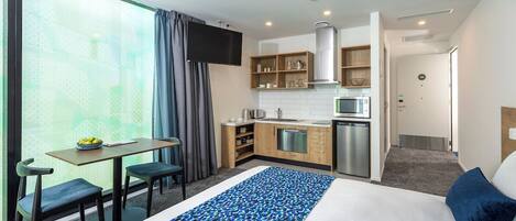 A modern hotel room with a large bed, a small kitchenette, a dining area, and a TV.