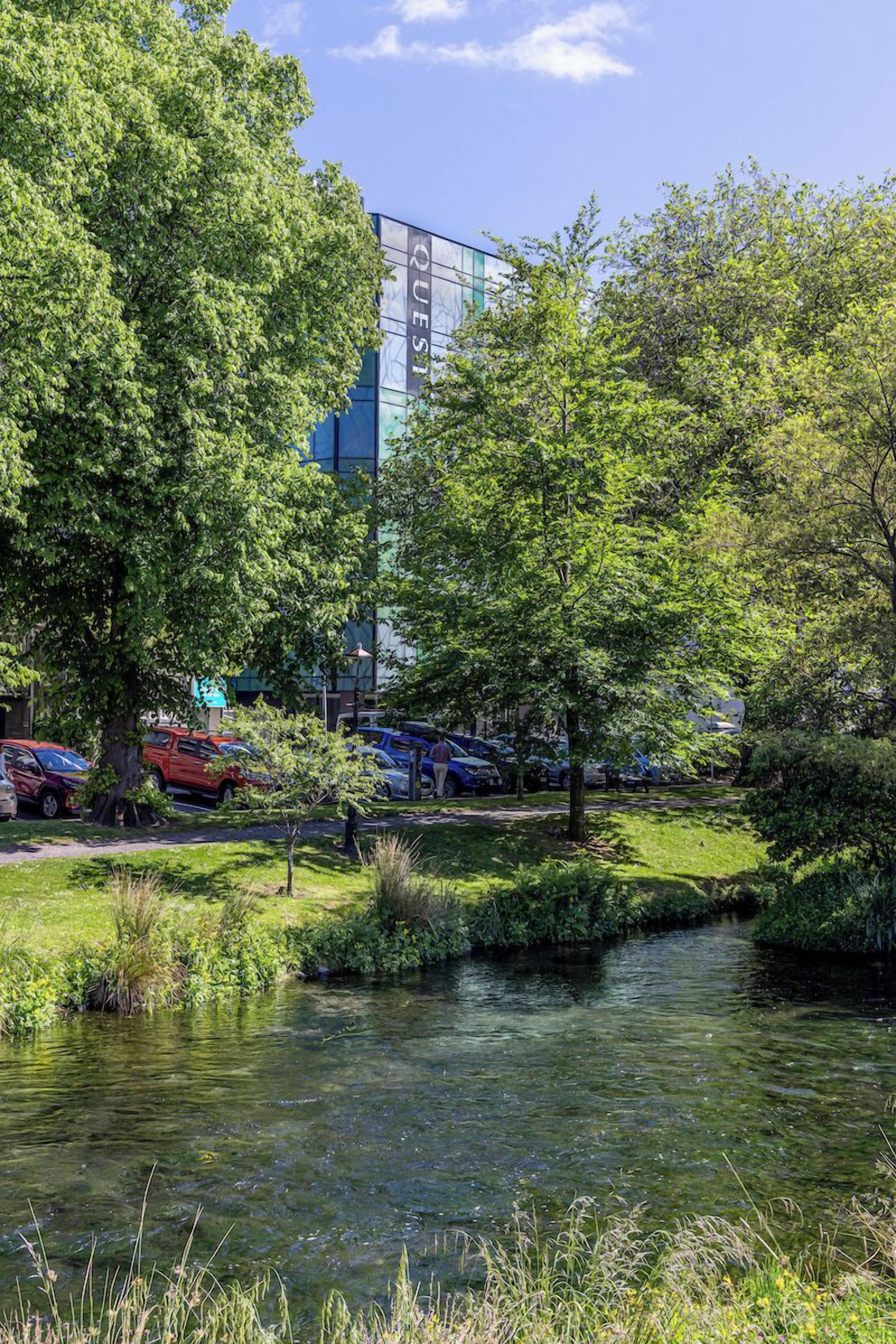 Un parc paisible avec un étang, une végétation luxuriante et un bâtiment moderne en arrière-plan.