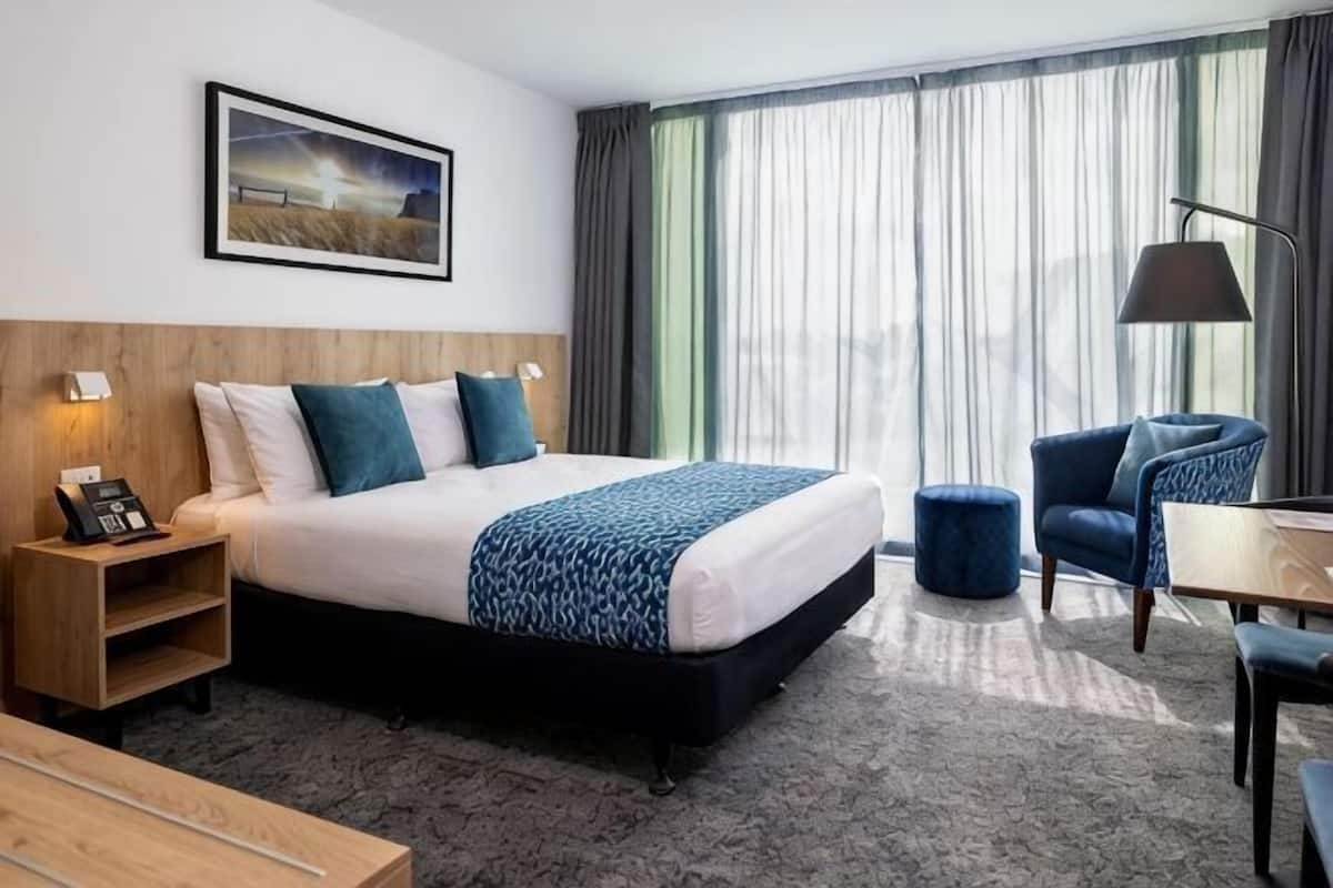 A hotel room with a bed, bedside table, lamp, chair, and a view of the outdoors through large windows.