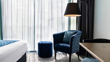 Ein Hotelzimmer mit einem Bett, einem blauen Sessel, einem runden Hocker, einem Schreibtisch und einer Lampe.