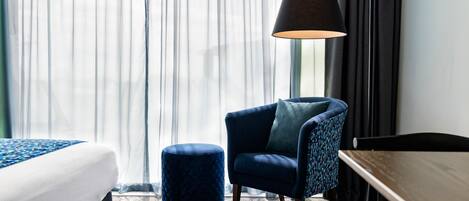 A hotel room with a bed, a blue armchair, a round ottoman, a desk, and a lamp.