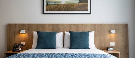 A hotel room with a large bed, two pillows, a bedside table with a phone, a wall-mounted light, and a framed picture of a sunset over a beach.
