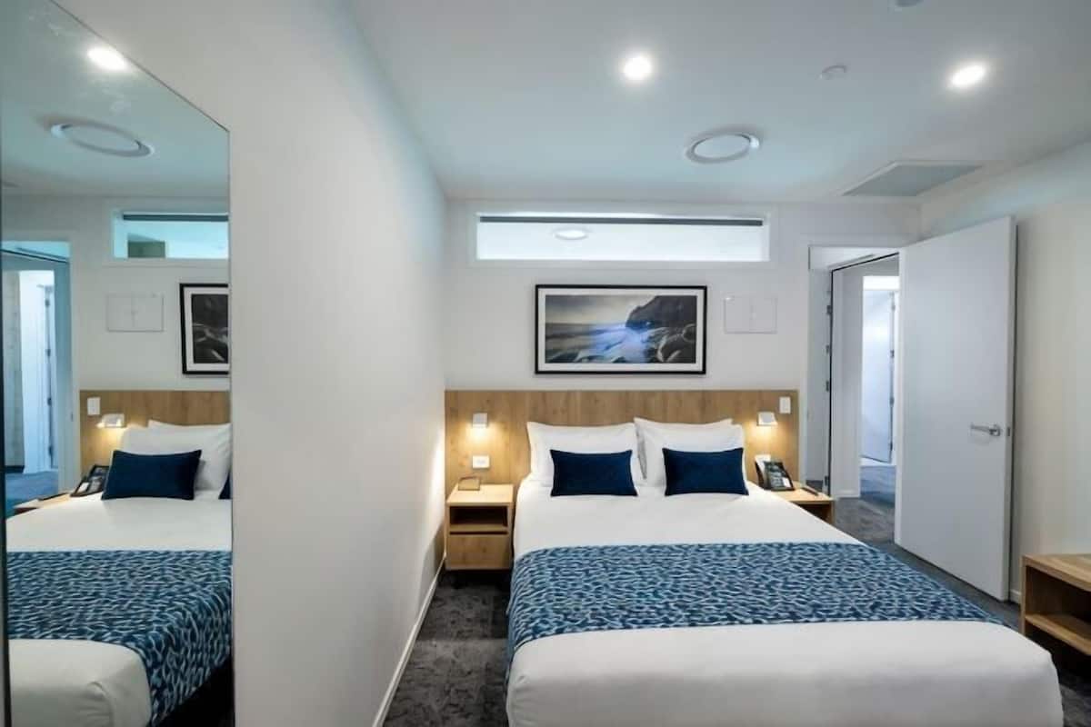 A hotel room with two beds, a large mirror, a nightstand with a lamp, a framed picture on the wall, and a door.