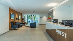 A reception area with a wooden counter labeled "QUEST," a seating area with sofas and chairs, and a view of the outdoors through large windows. - Quest on Cambridge  (Christchurch)