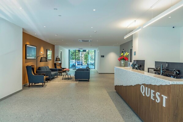 A reception area with a wooden counter labeled "QUEST," a seating area with sofas and chairs, and a view of the outdoors through large windows. - Quest on Cambridge  (Christchurch)