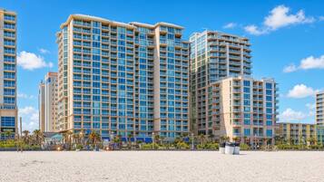 A high-rise hotel with multiple balconies, a sandy beach in the foreground, and palm trees.