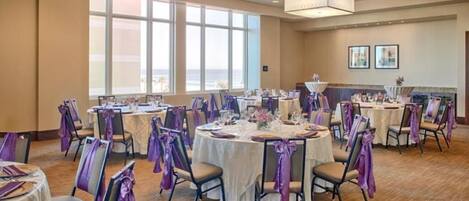 A banquet hall with round tables, chairs, and purple sashes, featuring large windows and ceiling lights.