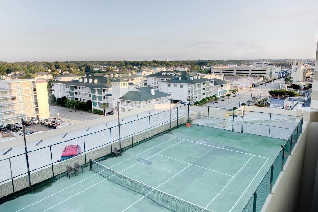 Una cancha de tenis con red, rodeada por una cerca, y contigua a un edificio con varios pisos y balcones.