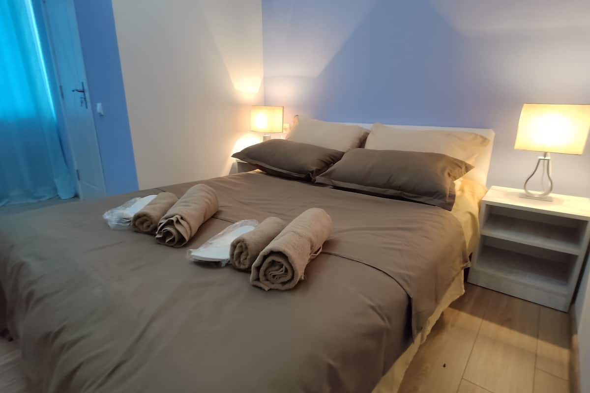 A neatly made bed with four rolled towels, a bedside table with a lamp, and a blue curtain in the background.