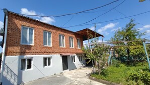 A two-story brick house with a red-tiled roof, a small porch, and a garden area with trees and plants.