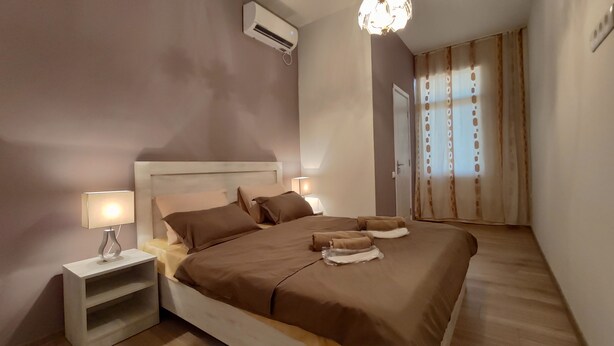 A bedroom with a large bed, two bedside tables with lamps, an air conditioning unit, and a window with curtains.