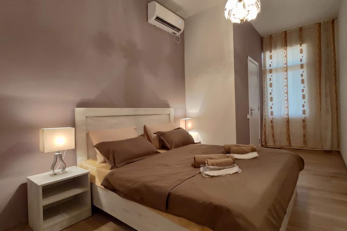 A bedroom with a large bed, two bedside tables with lamps, an air conditioning unit, and a window with curtains.