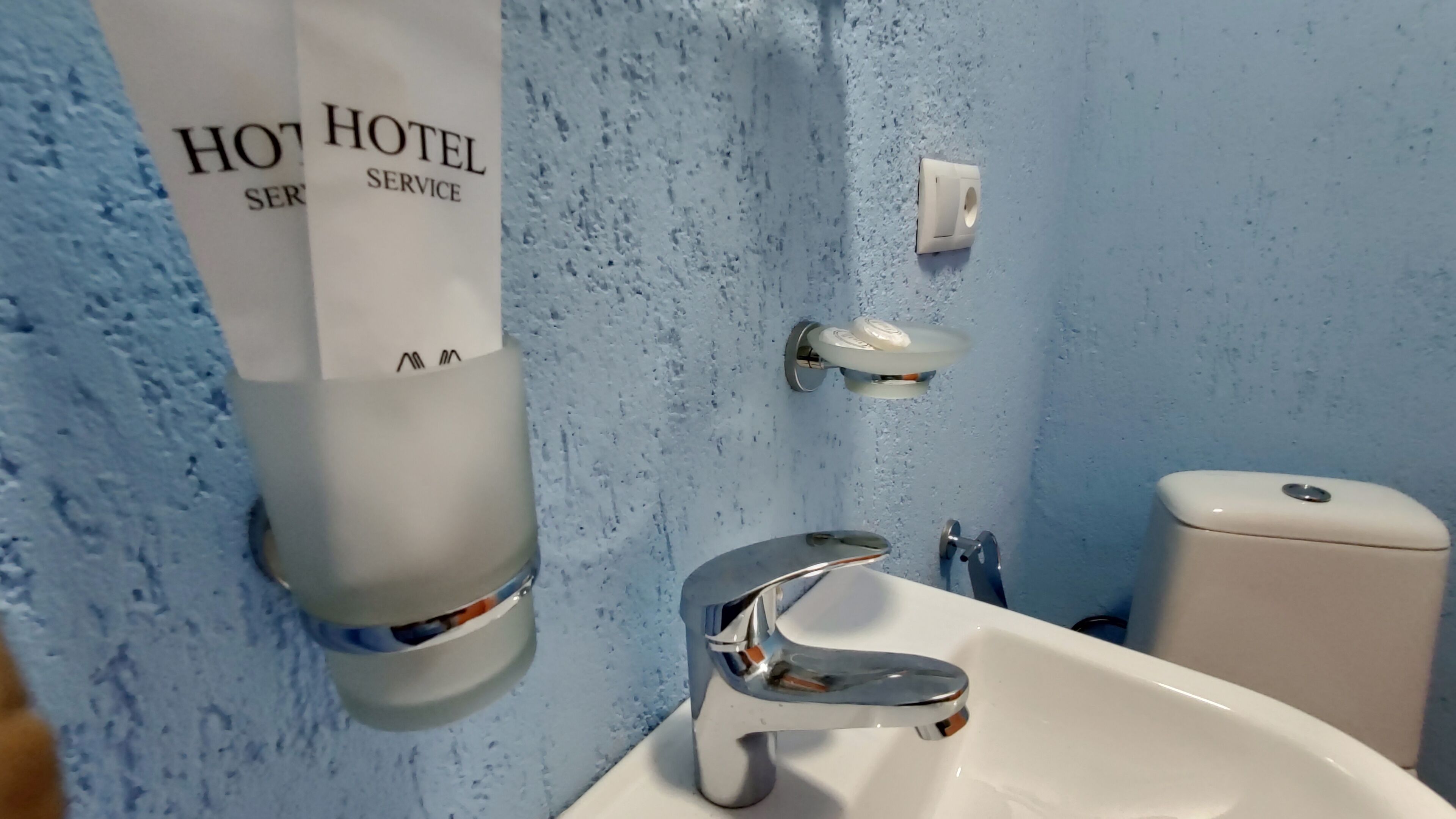 Un baño con un lavabo blanco, un espejo y un inodoro. Hay un cartel en la pared que dice 'HOTEL SERVICE'.