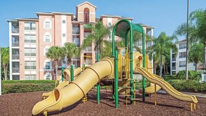 A playground with slides and climbing structures, surrounded by a landscaped area with palm trees and a multi-story building in the background.