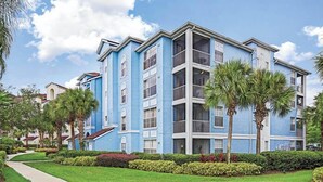 A multi-story hotel with a blue facade, balconies, and a well-maintained garden with palm trees and shrubs.