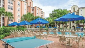 A pool area with lounge chairs and umbrellas, a ping pong table, and a building with multiple windows and balconies.