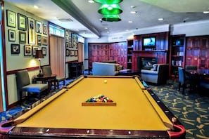 A pool table with a cue ball and a colorful rack of balls, surrounded by seating and a television in a room with framed pictures on the walls.