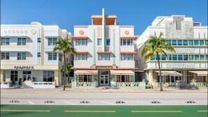 A mid-20th century hotel with a central entrance, palm trees, and adjacent shops.
