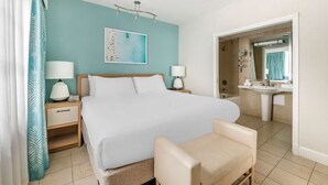 A hotel room with a bed, bedside lamps, a chair, a bathroom with a sink and mirror, and a painting on the wall.