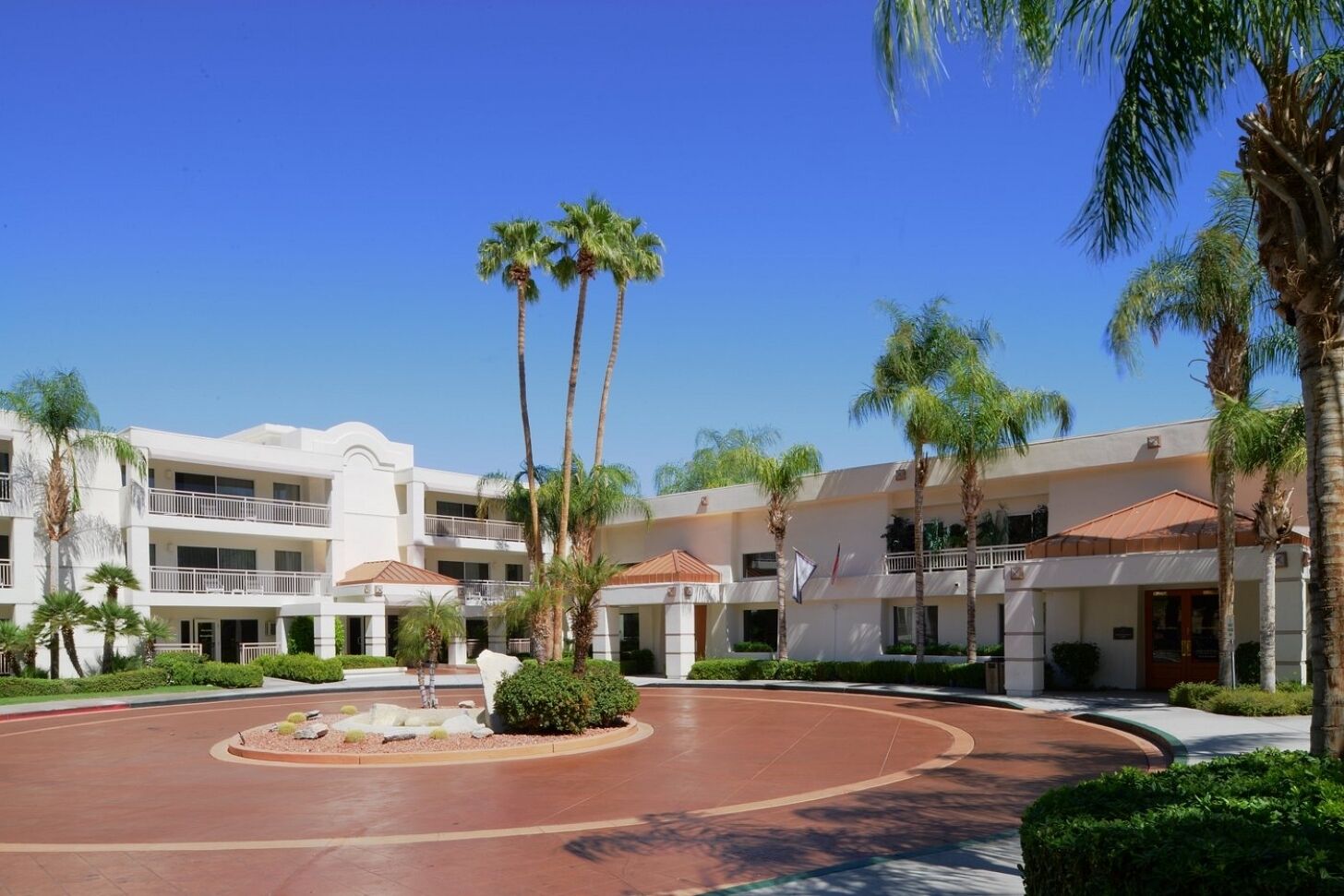 Un hotel de varios pisos con un patio central, palmeras y un cielo azul despejado.