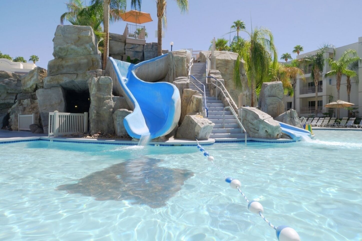 A pool with a water slide, stairs leading to a rock formation, and palm trees.