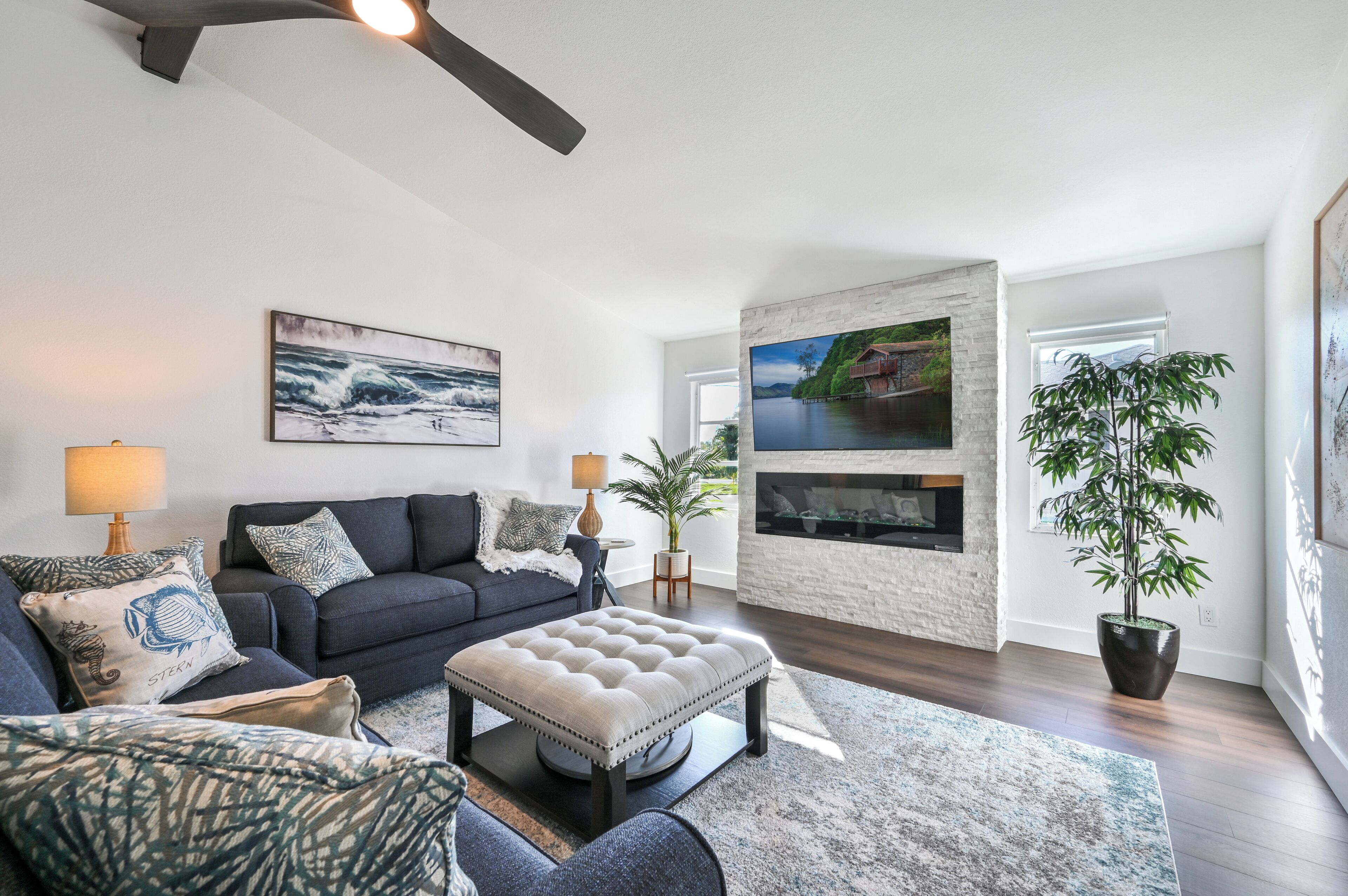 A modern living room with a dark grey sofa, a tufted ottoman, and a large window with a view of a house and greenery.