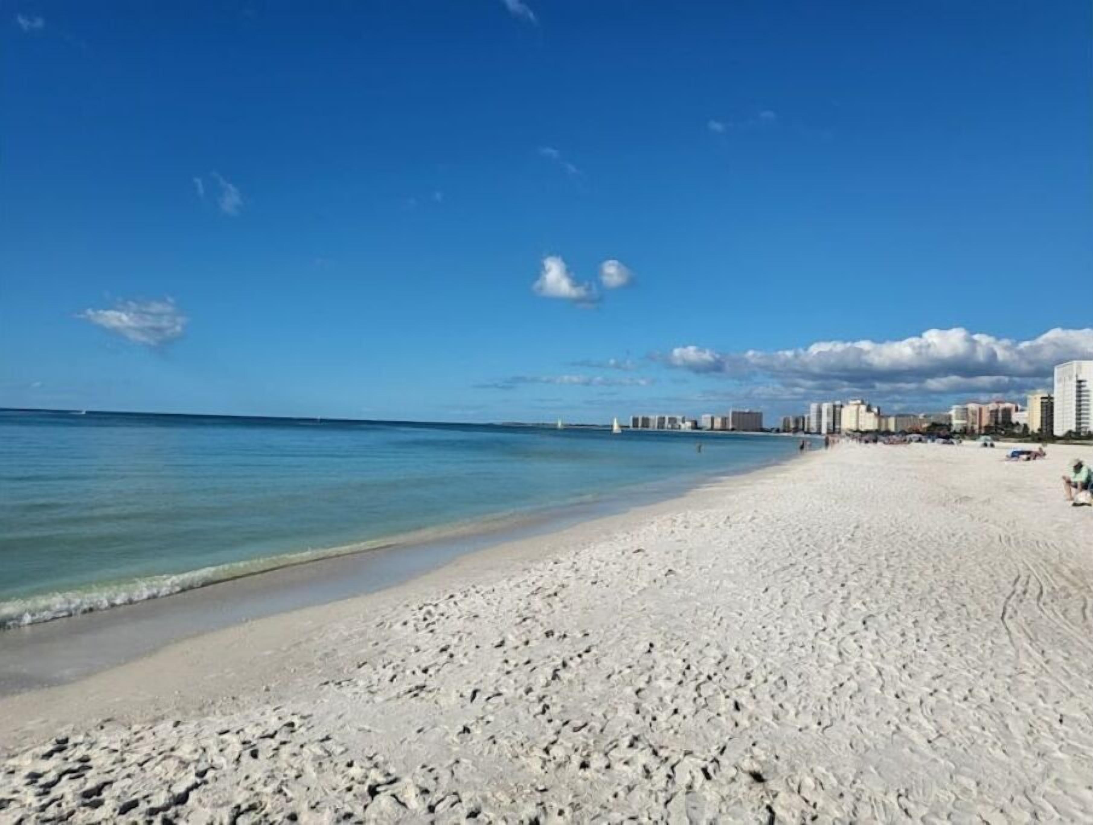Una extensa playa de arena con agua azul cristalina y un horizonte urbano lejano.
