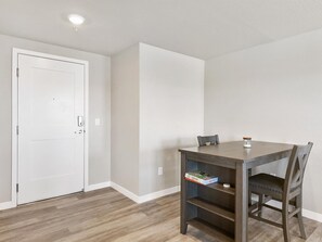 A room with a wooden floor, a white door, a table with a chair, and a small shelf with a book.