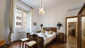 Une chambre à coucher comprenant un lit, deux tables de chevet, un luminaire suspendu, un radiateur, une fenêtre avec des rideaux et un plancher en bois.
