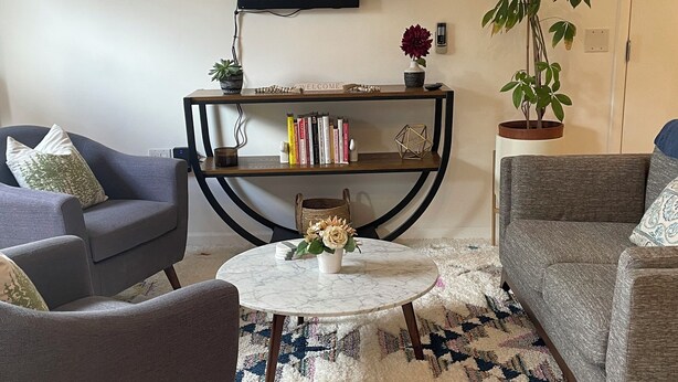 Uma sala de estar com uma TV de tela plana na parede, uma mesa de centro redonda com um vaso, dois sofás, uma estante de livros e um vaso de planta.