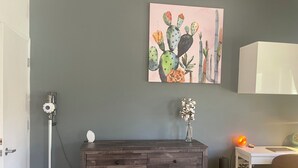 Quarto com penteadeira de madeira escura, pintura de cactos, vaso com flores secas, escrivaninha com luminária e uma cadeira.