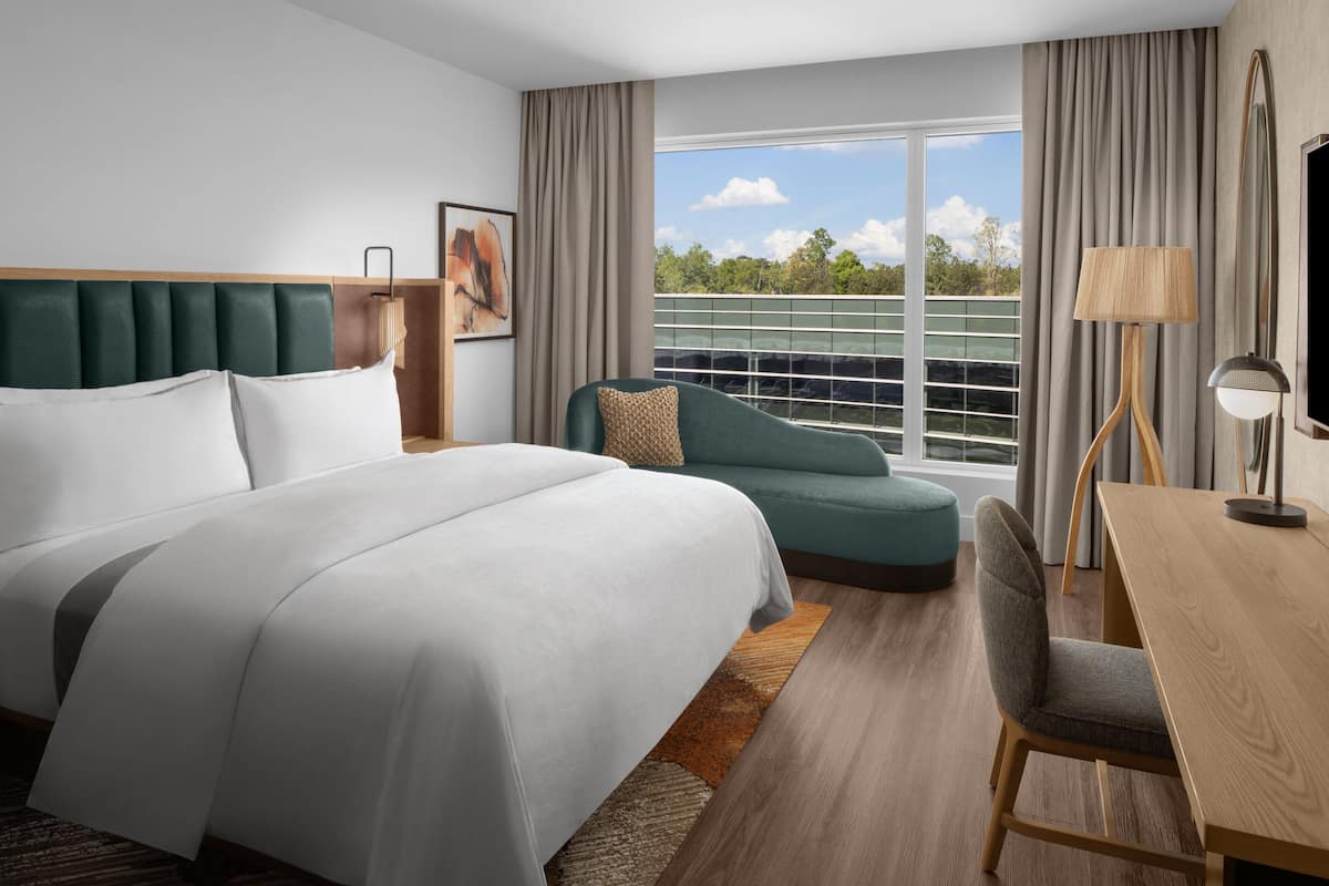 A modern hotel room with a large bed, a sofa, a dining table, and a view of the outdoors.