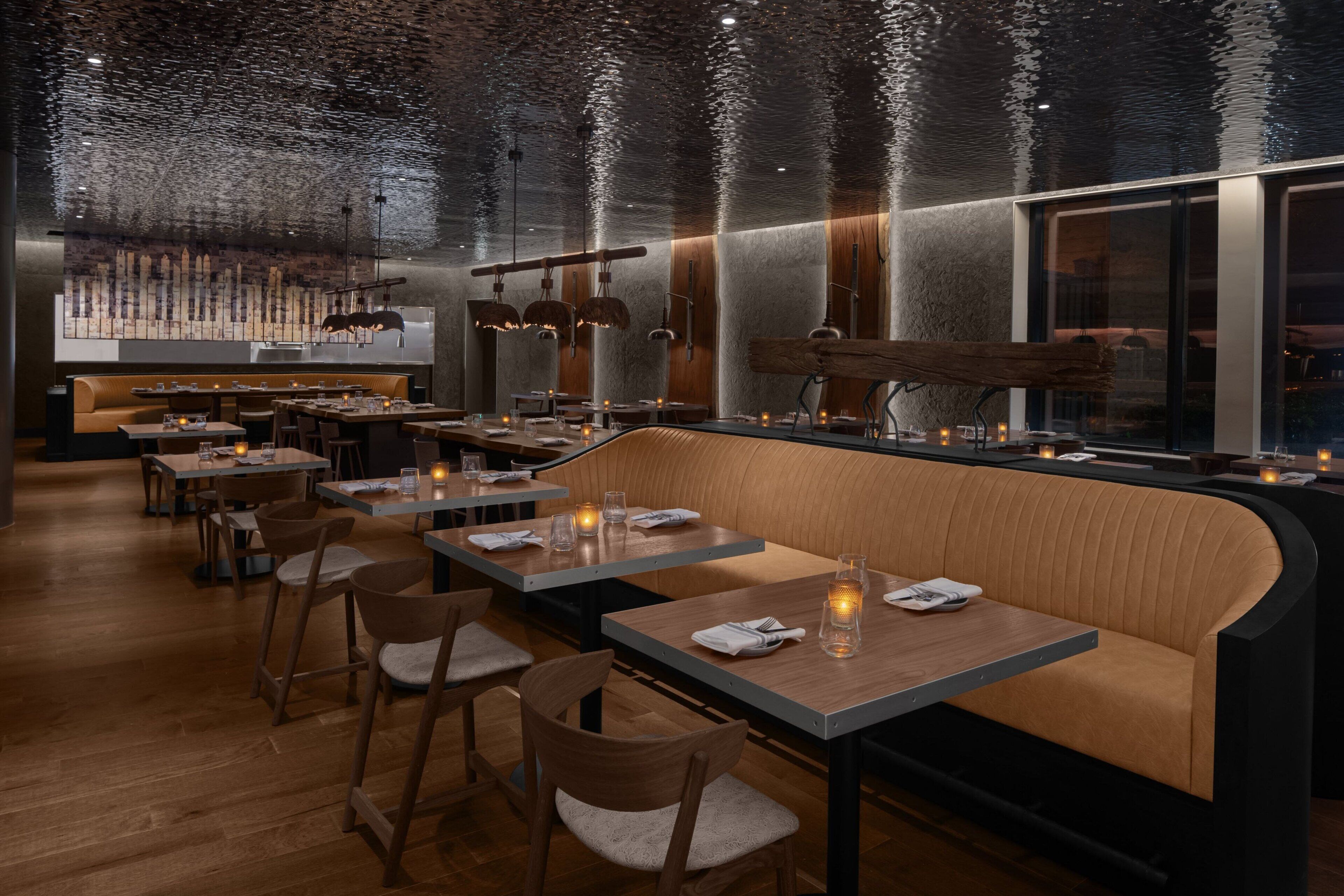 A modern restaurant with wooden tables and benches, ambient lighting, and a reflective ceiling.