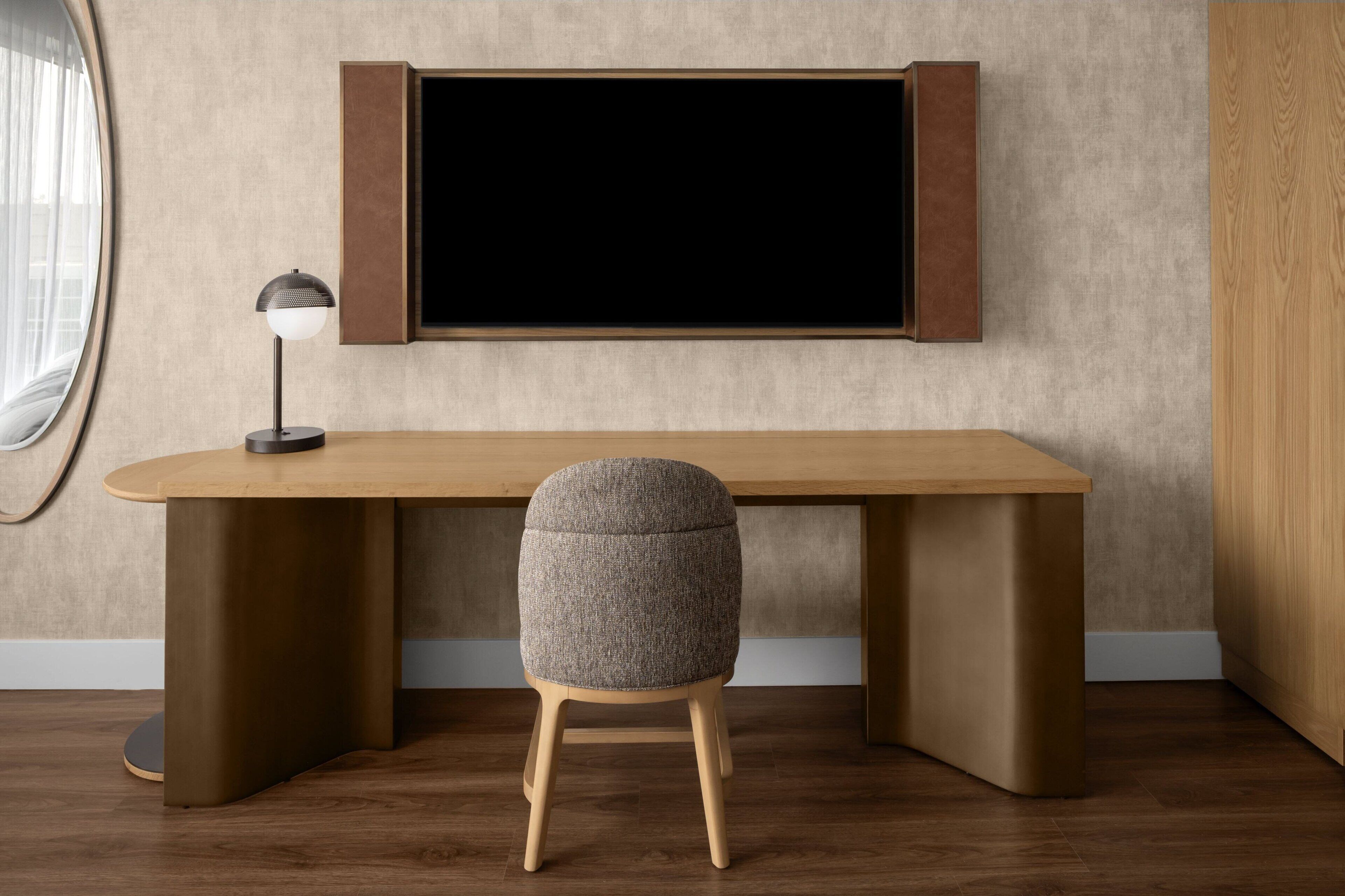 A modern room with a wooden desk, a grey upholstered stool, and a floor lamp.