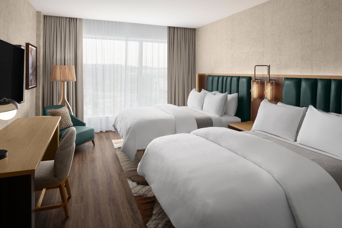 A hotel room with two beds, a desk, a chair, a TV, and a large window with curtains.