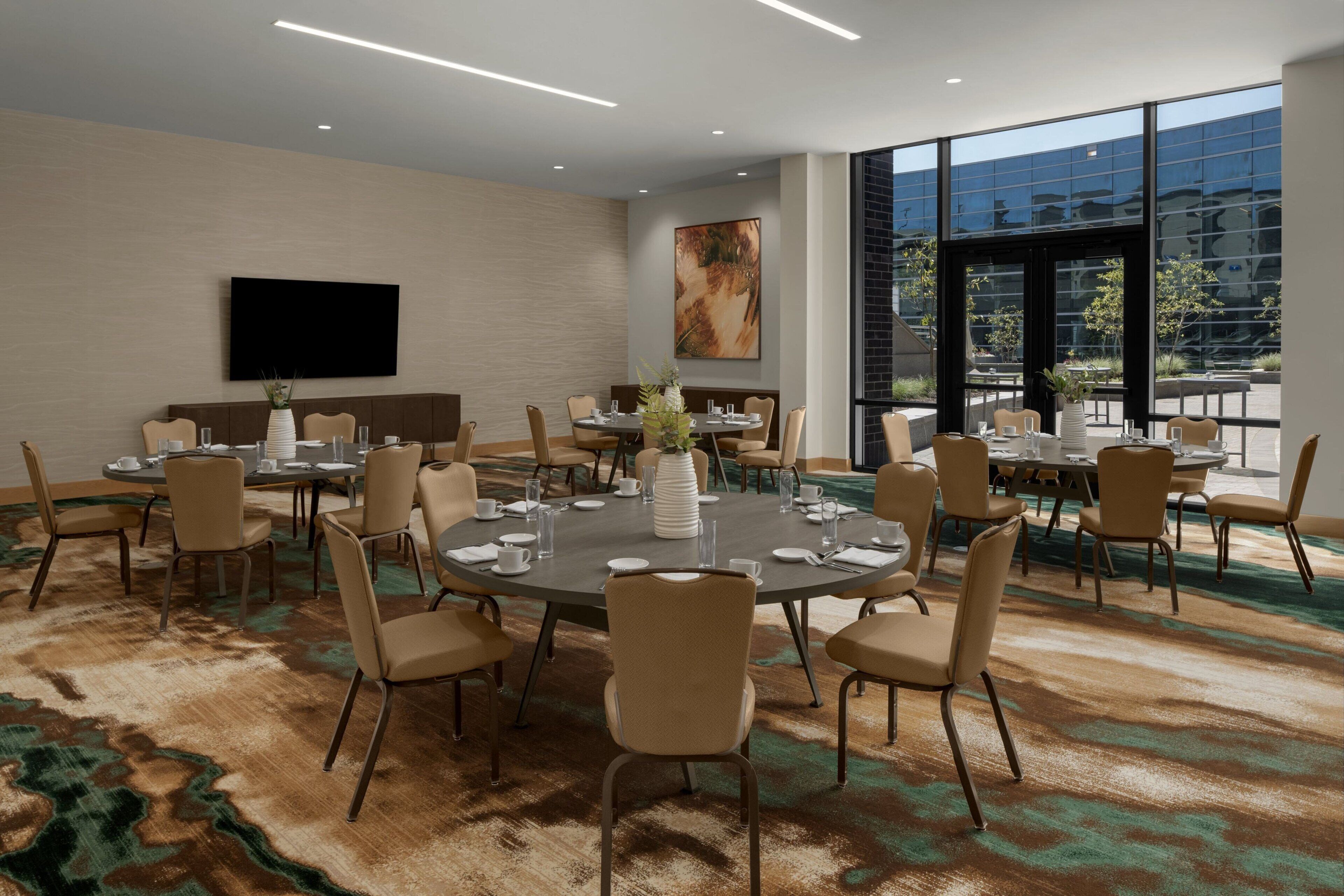 A conference room with round tables, beige chairs, a large window, and a mounted television.