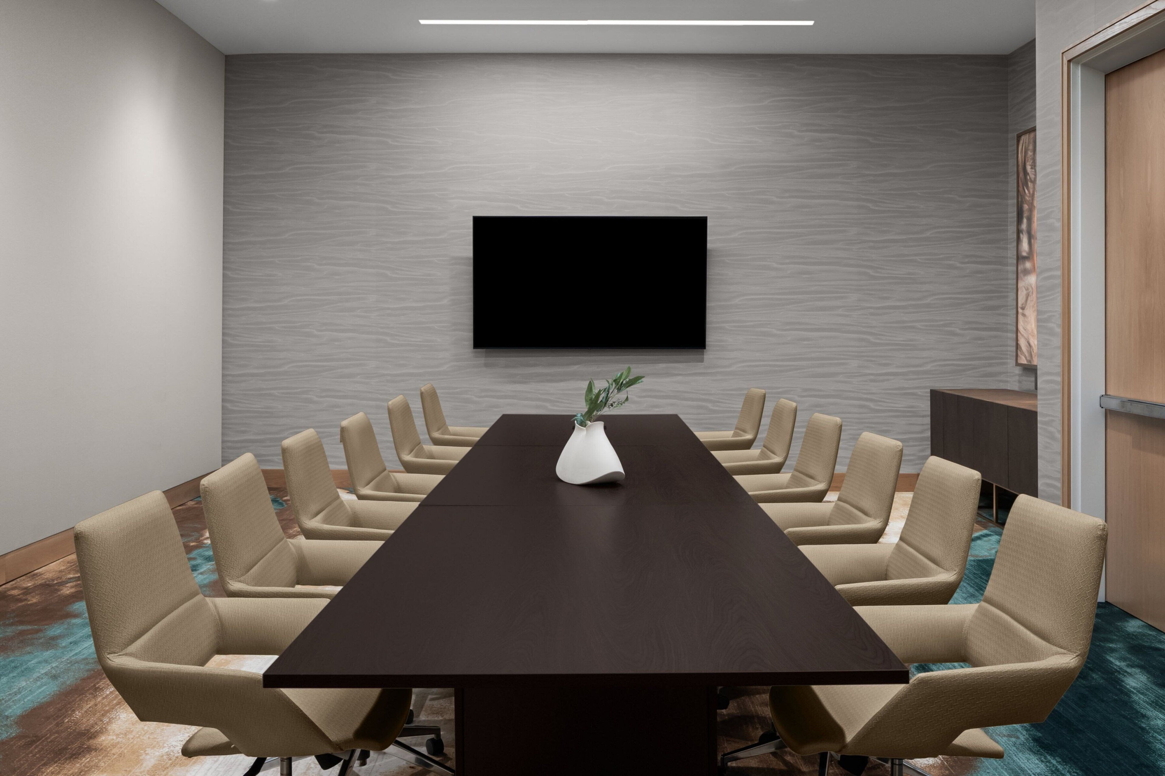 A conference room with a long table, beige chairs, a flat-screen TV on the wall, and a vase with a plant.