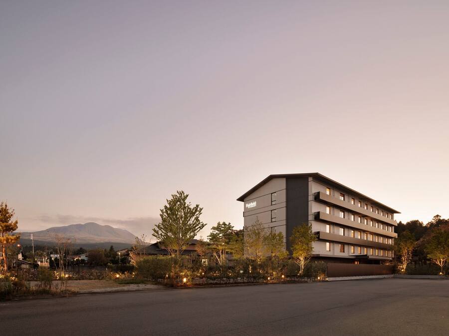 A modern hotel with a flat roof and multiple floors, surrounded by trees and a mountain in the background.