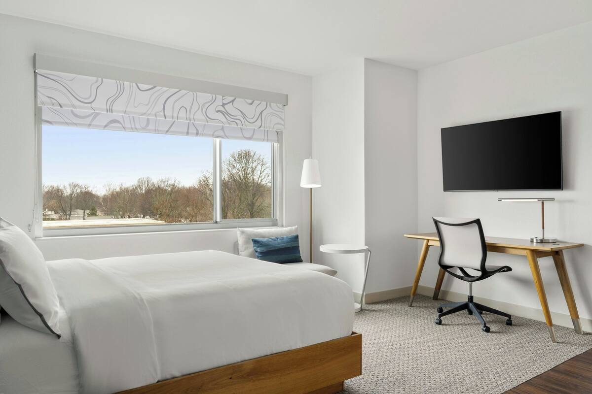 A modern hotel room with a large bed, a desk with a chair, a TV, and a window with a view of trees.