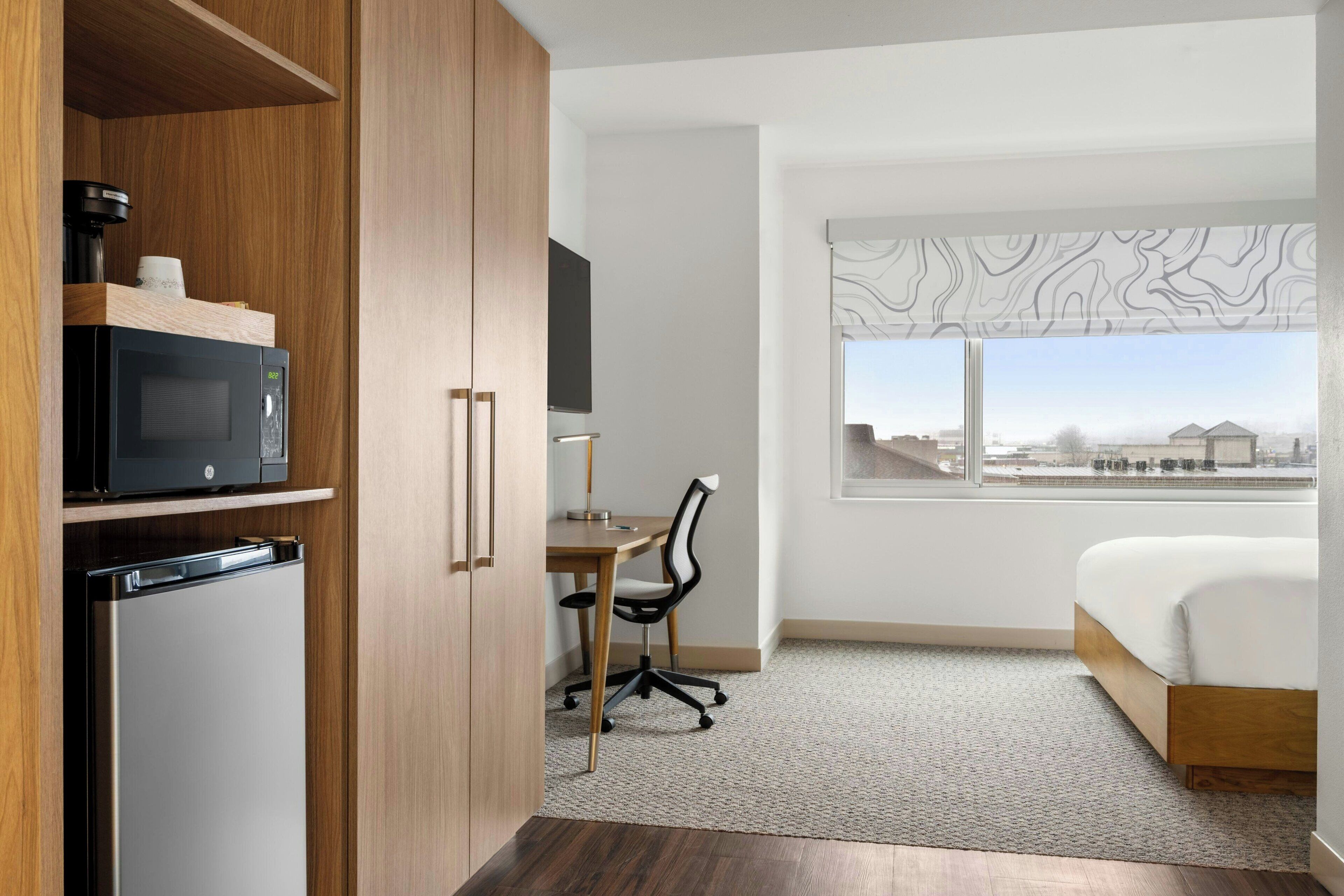 Ett hotellrum med en säng, ett skrivbord med en stol, en mikrovågsugn och ett kylskåp.