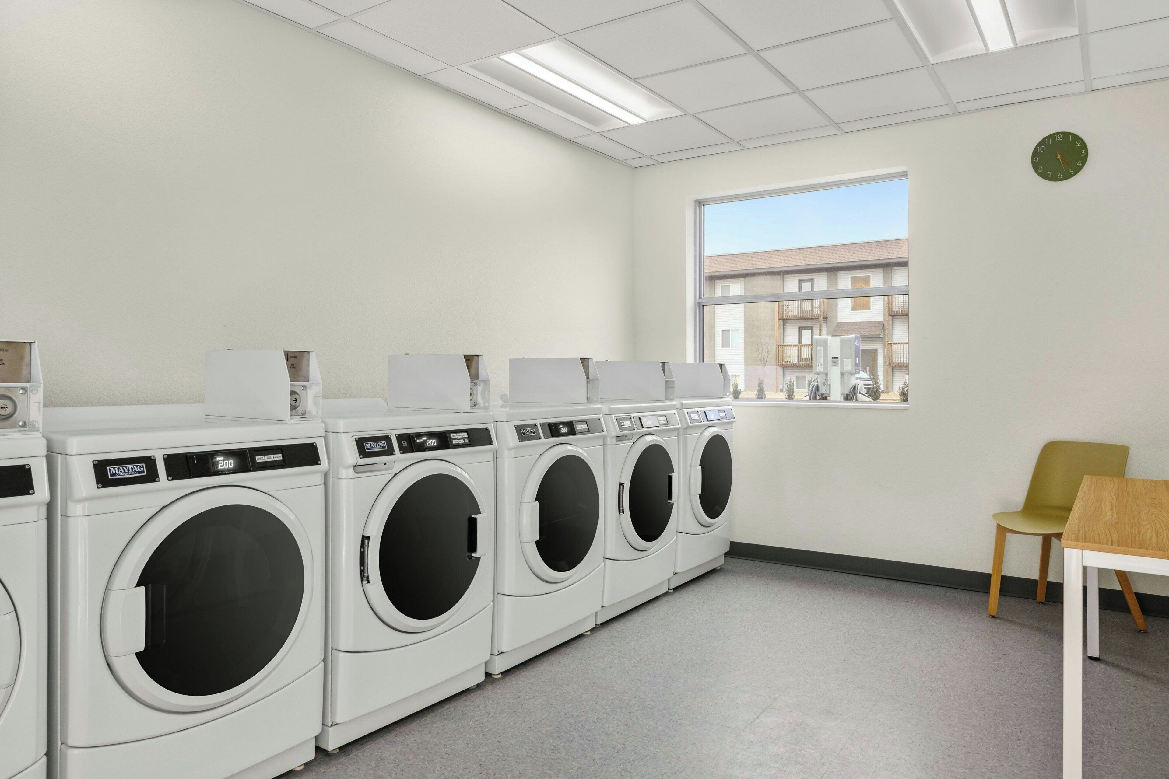 A laundry room with four white front-loading washing machines, a wooden table, and a window with a view of buildings outside.