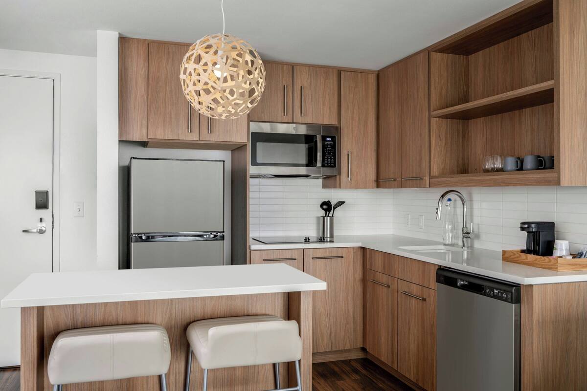 A modern kitchen with wooden cabinets, stainless steel appliances, a white countertop, and two bar stools.