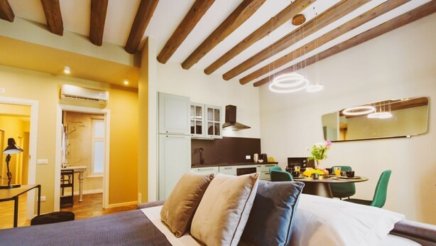 A bedroom with a bed, pillows, a desk, a chair, a mirror, and a kitchenette.