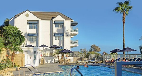 Enjoy a relaxing stay in a two-story hotel with a flat roof and balcony, surrounded by a pool area with lounge chairs and umbrellas.
