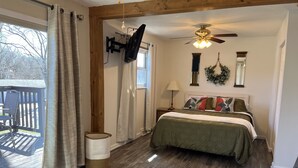 Une chambre à coucher comprenant un lit, un ventilateur de plafond, un téléviseur fixé au mur, une table de chevet avec une lampe, et une vue sur l’extérieur par une fenêtre.