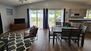 Een ruime woonkamer met een televisie, een bank, een eettafel met stoelen, een plafondventilator en uitzicht op de omgeving via grote schuifdeuren.