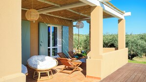 Een overdekt terras met rieten stoelen, een beklede bank, een houten terras en uitzicht op groen.